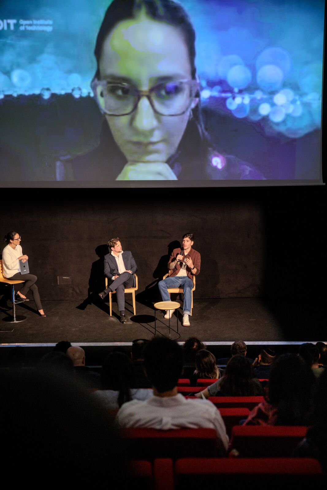On screen, Flatra Thaqi. On stage, from left to right: Greta Maiocchi, Daniel Graditi and Cristobal Bragagnolo